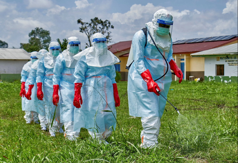 Team of workers in full PPE (blue suits, face shields, red gloves) spraying a field with a disinfection device outside a rural clinic.