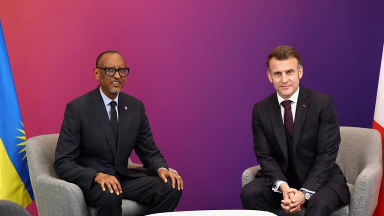 Two men in dark suits sit in gray chairs before a purple gradient backdrop, with a flag on each side.