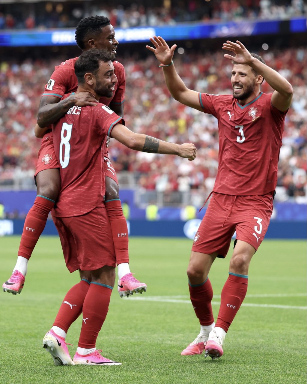 Three Portugal national team players in red jerseys celebrate a goal on the field, arms raised and smiling as a crowd cheers in the background.