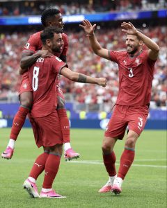 Three Portugal national team players in red jerseys celebrate a goal on the field, arms raised and smiling as a crowd cheers in the background.