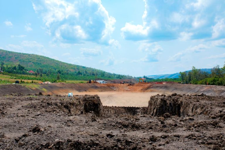 Open-pit construction site with a large crater, dirt piles, and distant machinery against a hilly, blue sky backdrop.