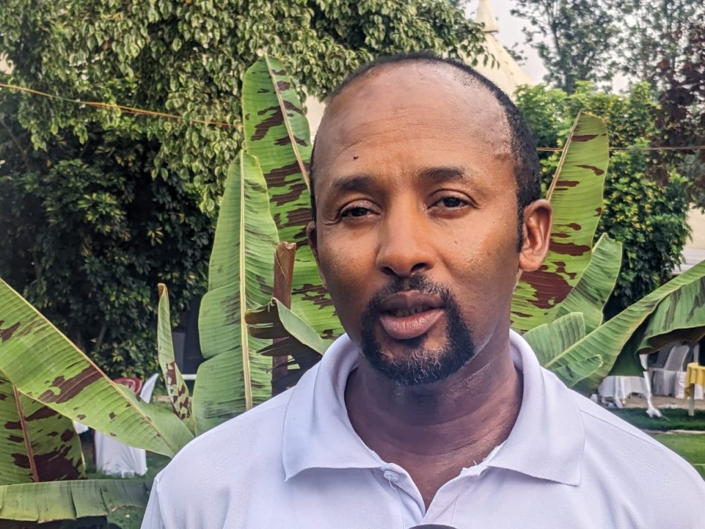 Man outdoors wearing a light gray polo shirt with large banana leaves and greenery in the background, looking at the camera.