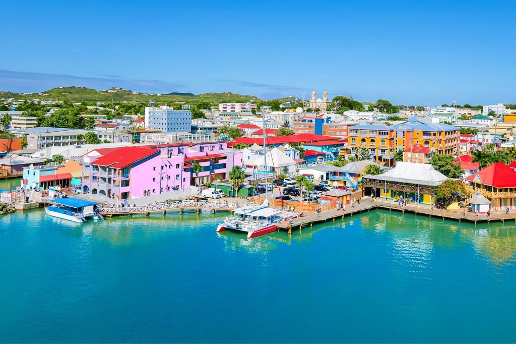 Colorful harbor town with pastel buildings along the water, boats docked at a wooden pier under a clear blue sky.
