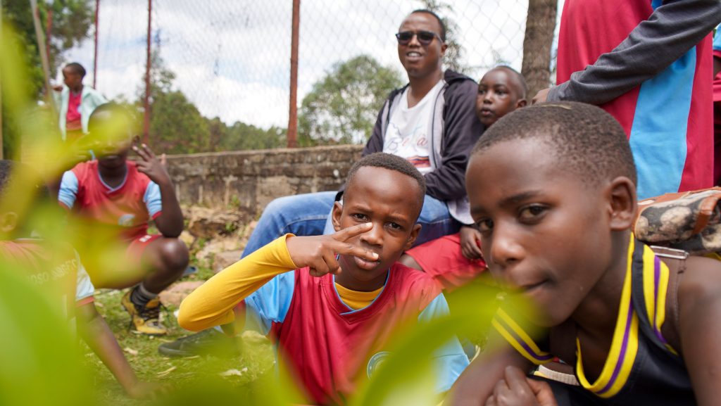 Group of kids in colorful sports jerseys posing for the camera outdoors near a chain-link fence.