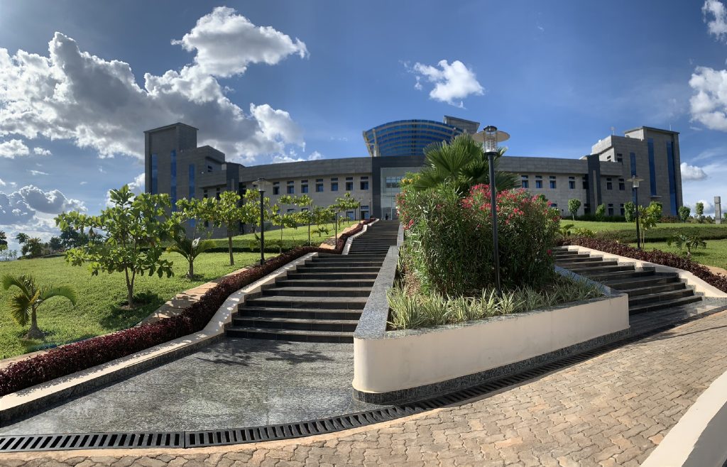 Modern institutional building on a landscaped hill with stairs and low hedges under a blue sky with clouds.