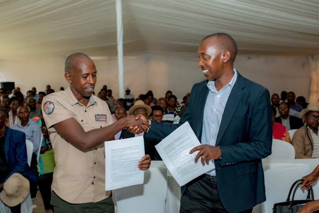 Two men shake hands and exchange documents at a formal event inside a large tent, with seated attendees in the background.