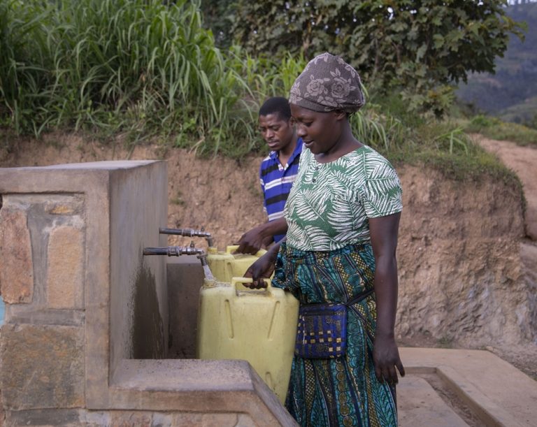 Woman in patterned dress fills a yellow water container at an outdoor hand pump with a man nearby in a rural setting