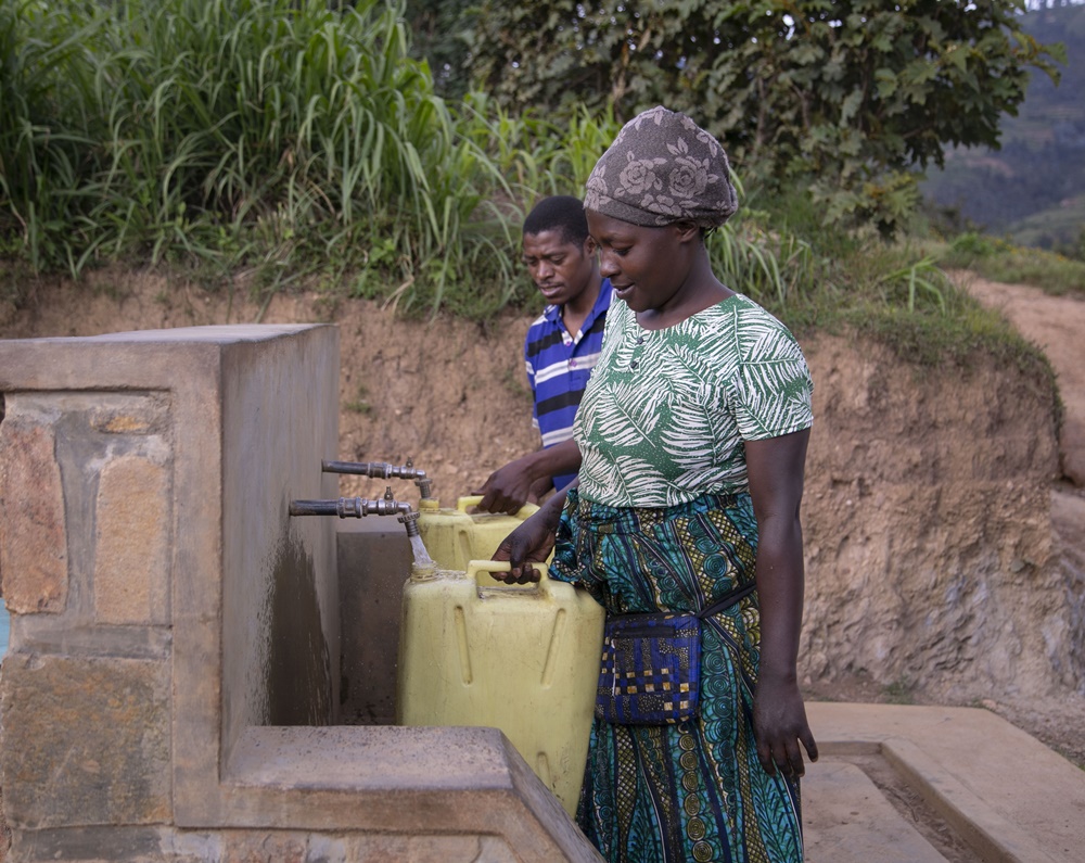 Woman in patterned dress fills a yellow water container at an outdoor hand pump with a man nearby in a rural setting