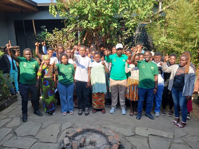 Group of about 30 people posing outdoors in a courtyard, many in green shirts, giving thumbs up for a team/community photo.