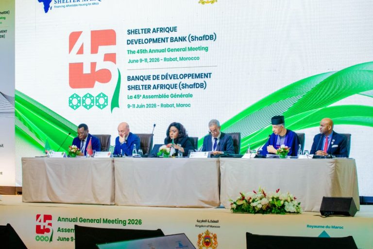 Panel of diverse speakers seated at a long table on stage during a conference, with a large screen behind showing '45' and AGM details in green and blue graphics.