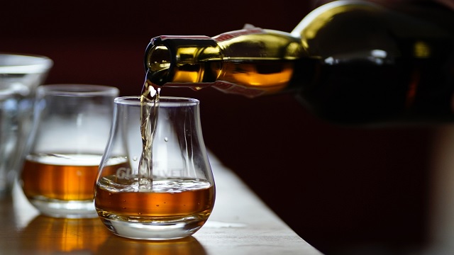 Whiskey being poured from a bottle into a short glass on a wooden bar with amber liquid mid-pour