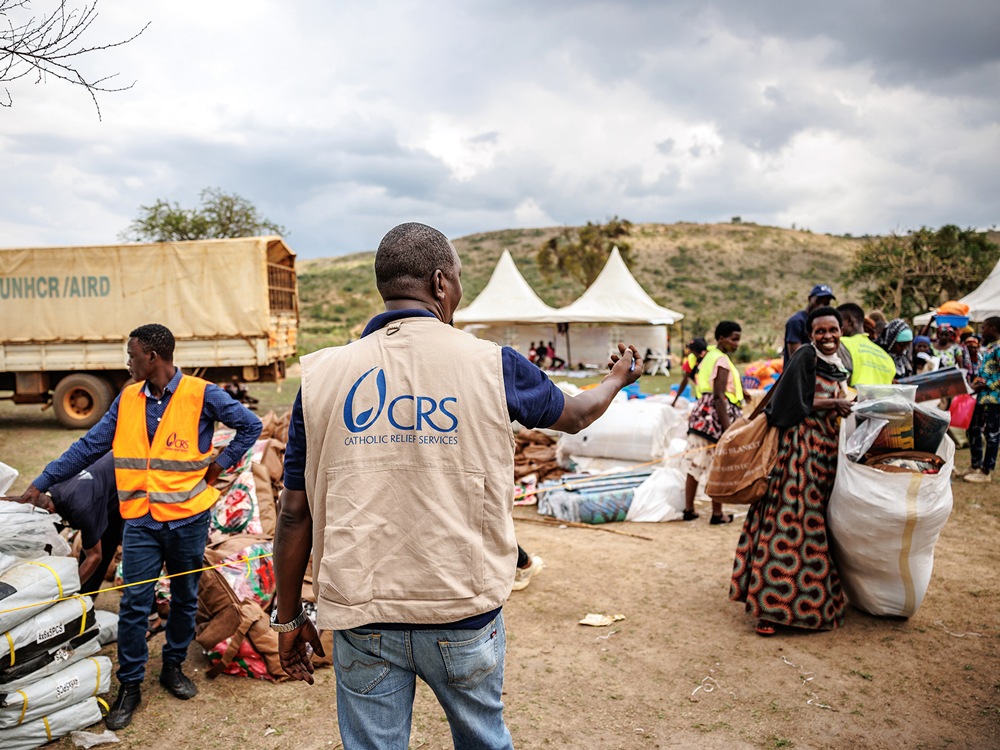 Aid workers from Catholic Relief Services distribute relief supplies to people in an open field with tents in the background and a truck nearby in a humanitarian aid setting.