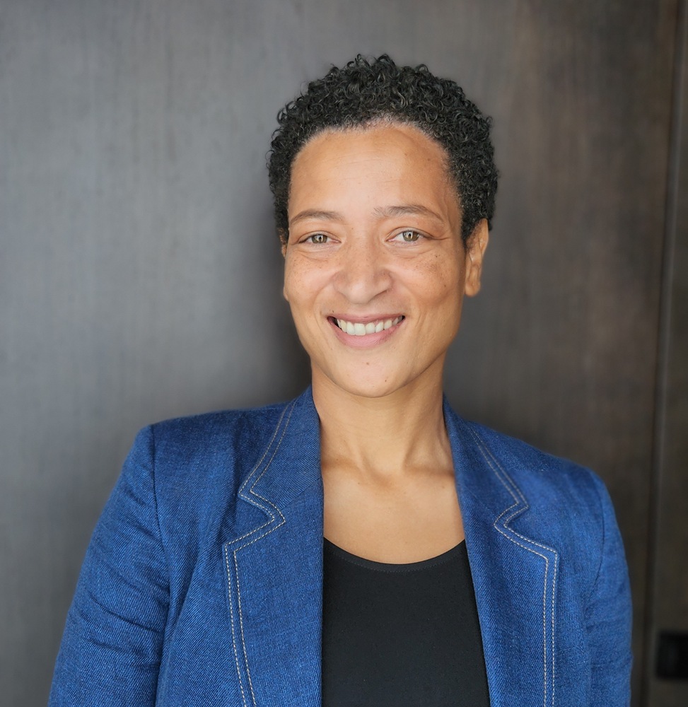 Professional headshot of a smiling woman with short curly hair wearing a blue blazer against a dark background.