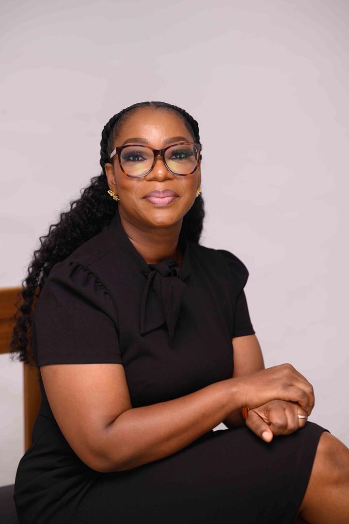 Portrait of a Black woman wearing large tortoiseshell glasses and a black dress, seated with hands folded in her lap.