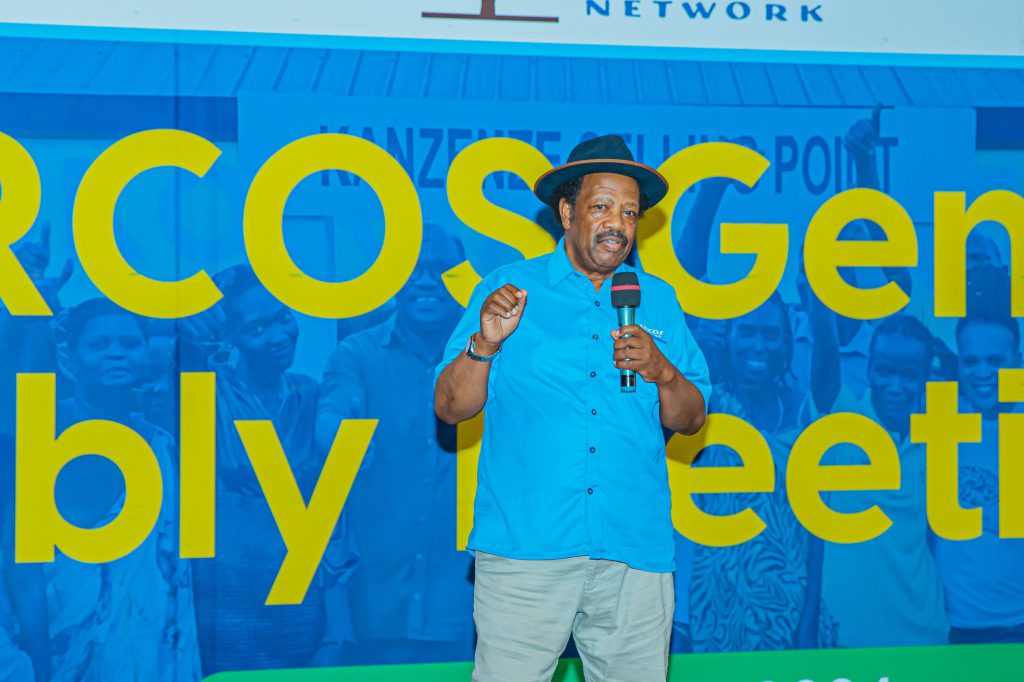 A man in a blue shirt and hat speaks into a microphone on a stage, with a blue banner featuring oversized yellow text behind him.