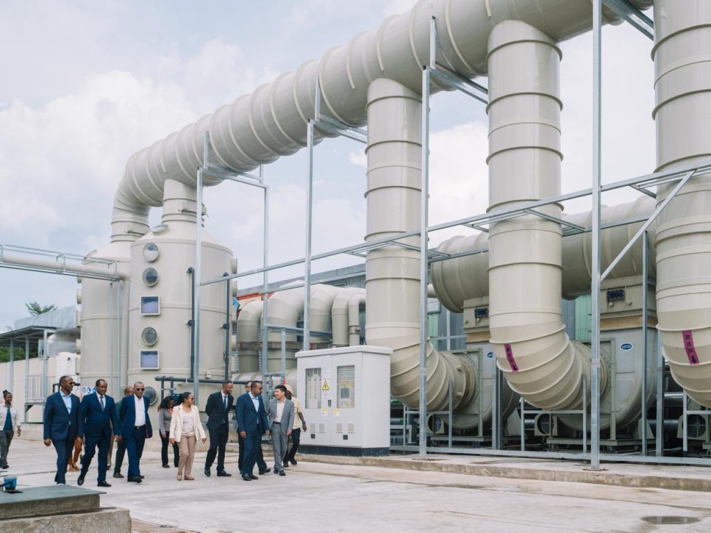 Group of professionals in suits walking among large beige industrial pipes and equipment at a plant.