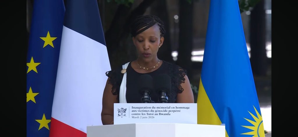 Speaker at a podium during a memorial ceremony with France, EU, and Rwanda flags in view behind her.