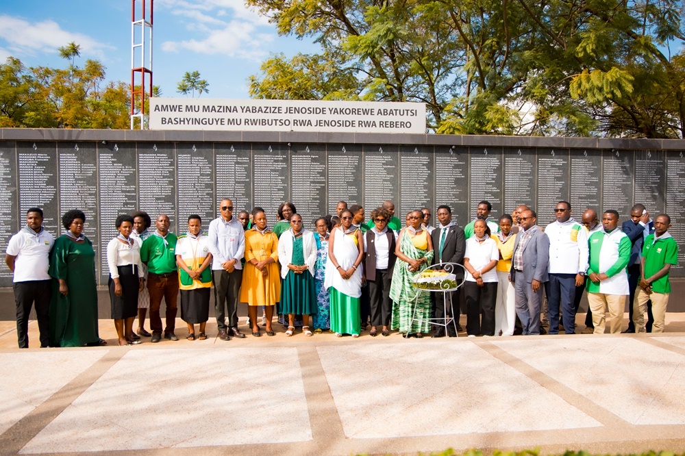Group of men and women in colorful attire posing in front of a memorial wall listing names on a sunny day.