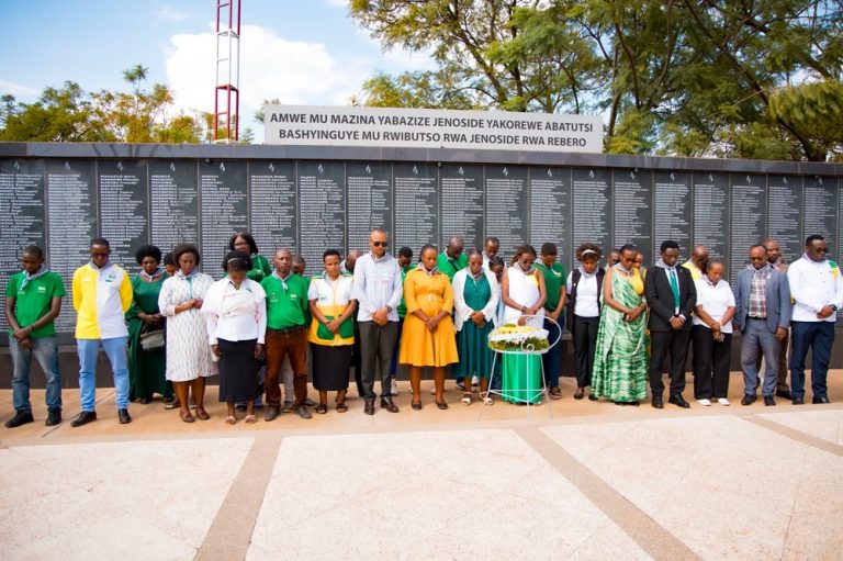 Diverse group of men and women standing in a line in front of a long memorial wall covered with names, outdoors with a wreath nearby.
