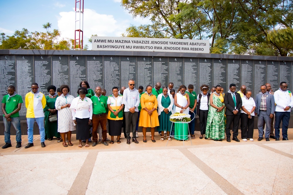 Diverse group of men and women standing in a line in front of a long memorial wall covered with names, outdoors with a wreath nearby.