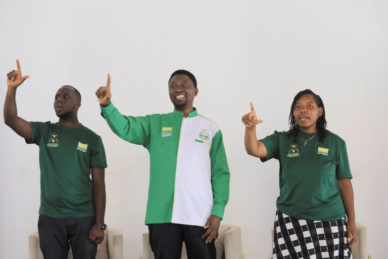 Three people stand side by side, each raising one finger and smiling, wearing green shirts with flag patches against a light background.