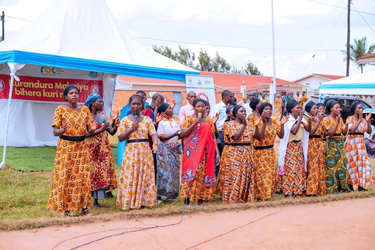 Group of women in colorful patterned dresses singing outdoors with a microphone under a white tent.