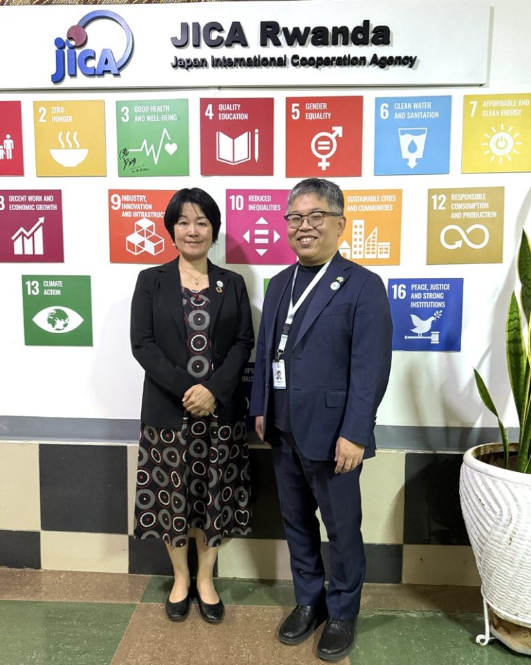 Two professionals stand in front of a JICA Rwanda backdrop with colorful SDG posters, smiling at the camera.