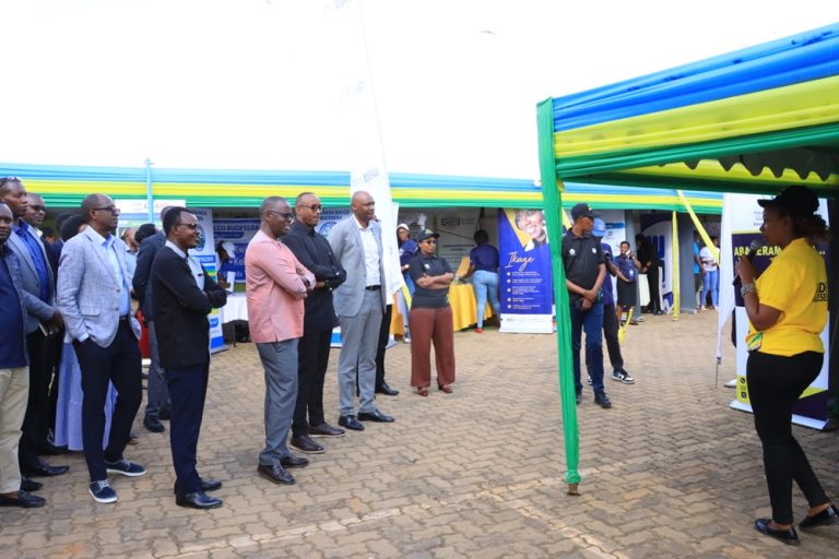 Group of professionals in business attire listening to a speaker at an outdoor expo under blue‑green canopies with promotional banners nearby