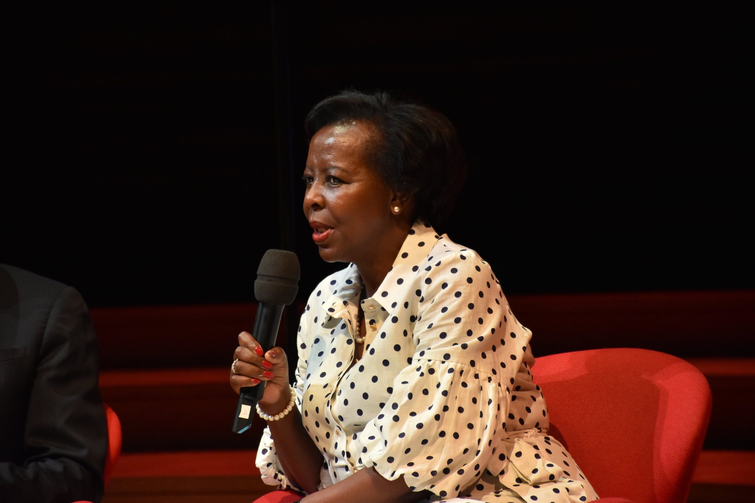 Woman in a cream polka-dot blouse speaks into a handheld microphone on a dark stage.