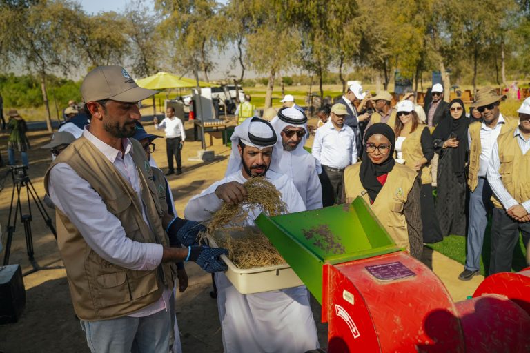 Group of people in outdoor event watching a composting demonstration with a man in traditional white dress holding plant material near a green chute and red bins.