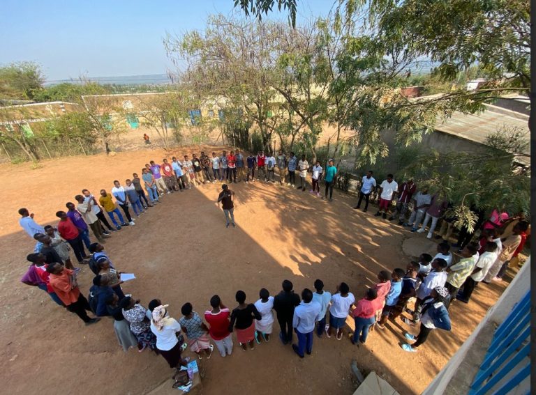 A large group of people arranged in a circle in a dusty outdoor yard, with a person speaking in the center under a tree on a sunny day.