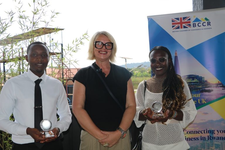 Three people posing at an awards event: a man in a white shirt and black tie on the left, a smiling woman in black with glasses in the middle, and a woman in white holding a trophy on the right, with a BCCR banner in the background.