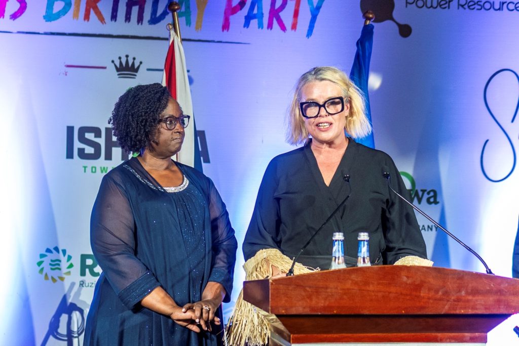 Two women at a wooden podium on a brightly lit stage; one speaks into a microphone while the other stands nearby, with flags and banners in the background.