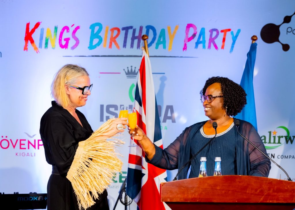 Two women toast with champagne flutes on a stage at a birthday party, colorful banner reading 'KINGS BIRTHDAY PARTY' behind them.