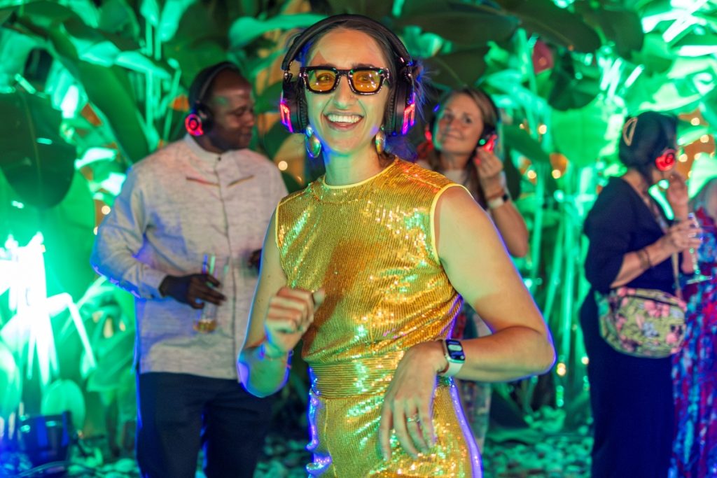 Woman in a gold sequined dress dancing with colorful headphones at a neon-lit party, smiling at the camera