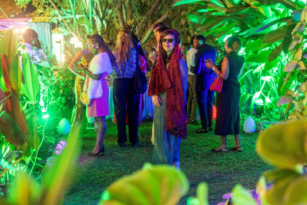 Group of people at a garden party under colorful lights; a woman in the center wears sunglasses and a red patterned scarf. John? (context-friendly) Not needed.