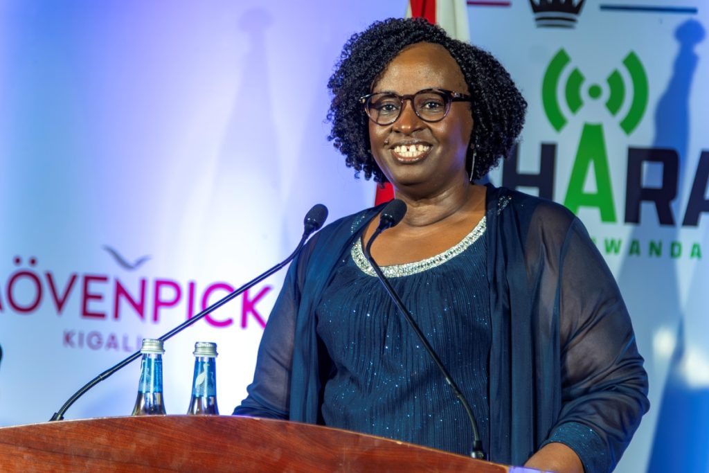 Smiling woman with glasses speaking at a podium with two microphones, banners in the background.