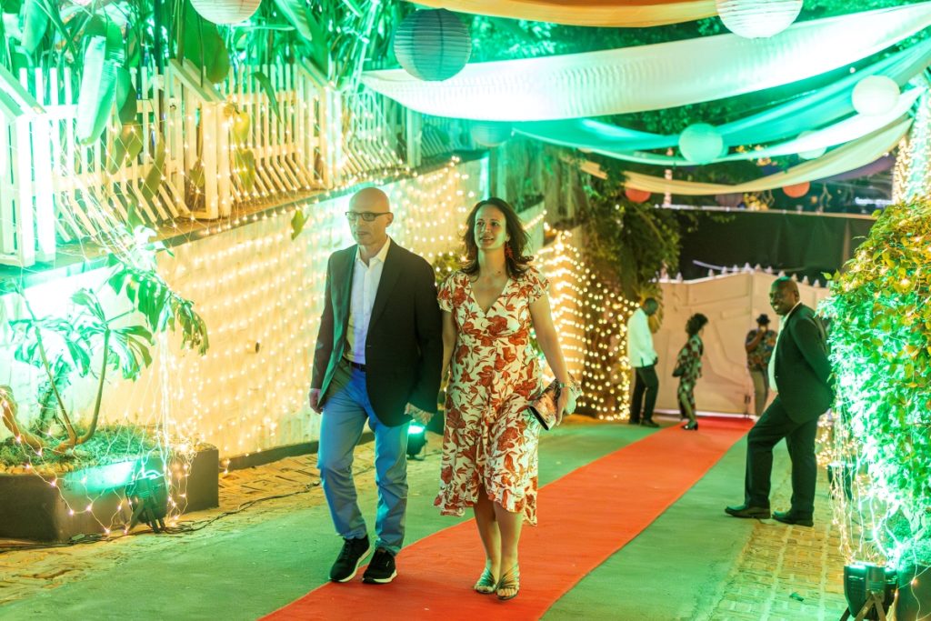 A couple walks a red carpet at an evening event, surrounded by warm fairy lights and green drapery.