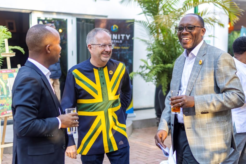 Three men in suits chatting and laughing at an outdoor event, one wearing a bold Jamaican flag-inspired shirt and holding a glass of drink.