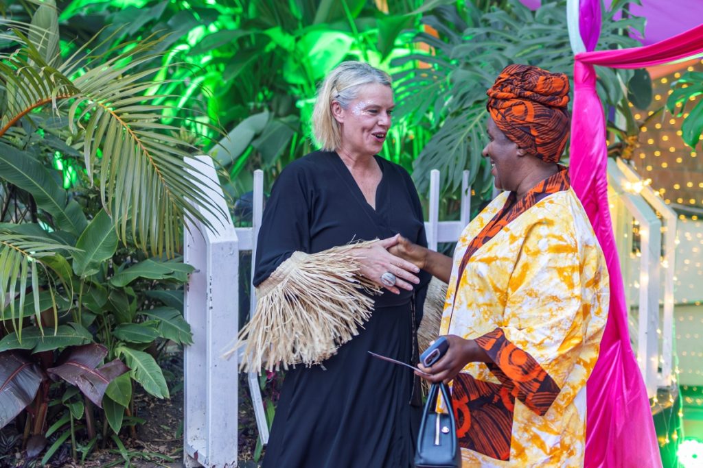 Two women share a friendly chat at an outdoor tropical-themed event; one in a black dress with fringe sleeves, the other in a bright yellow-orange patterned outfit and orange headwrap, with pink drapery and string lights behind them.