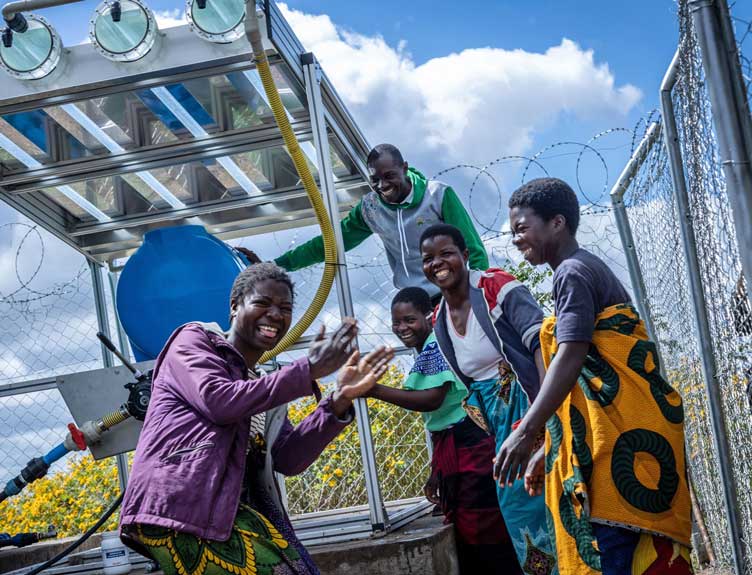 Six people celebrate around a solar-powered water pump installation outdoors, smiling and giving high fives.