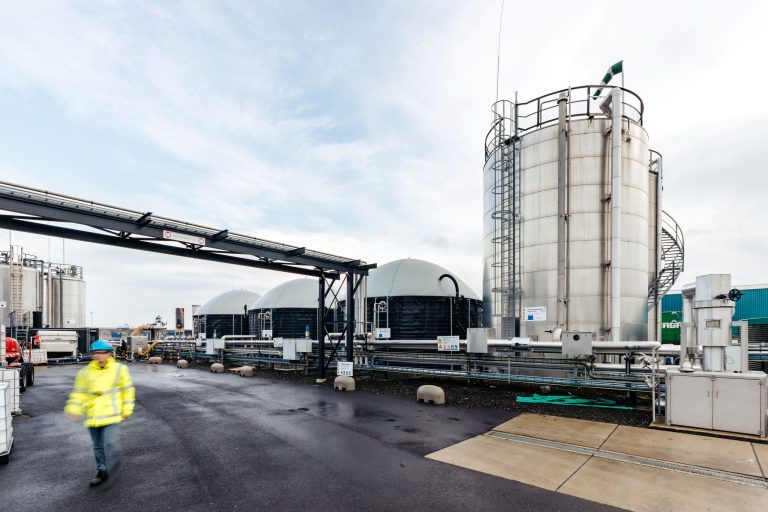 Industrial facility with tall stainless steel storage tanks, pipes, and a worker in a yellow safety jacket in the foreground.