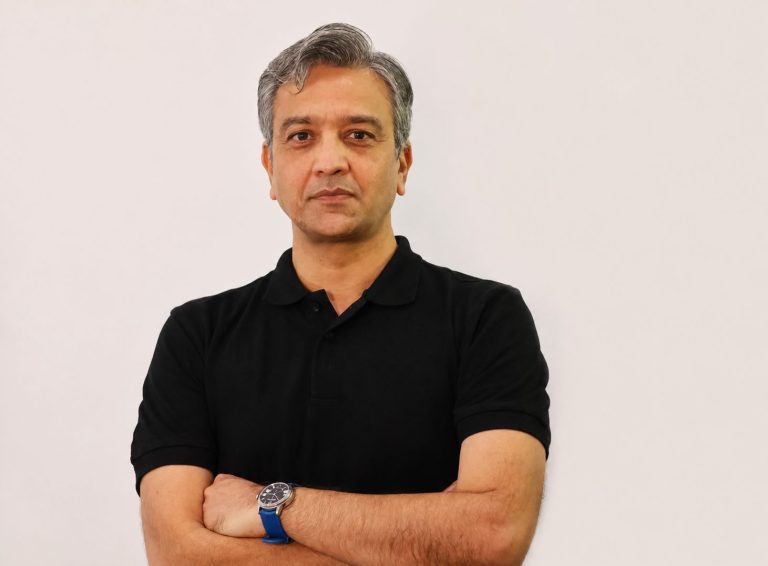Portrait of a middle-aged man with gray hair, wearing a black polo and a blue watch, arms crossed against a light background.