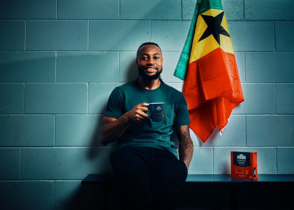 Man sits on a bench by a teal brick wall, holding a mug; a Jamaican flag hangs beside him and a box of Ahmad Tea London Blend rests on the bench beside him.