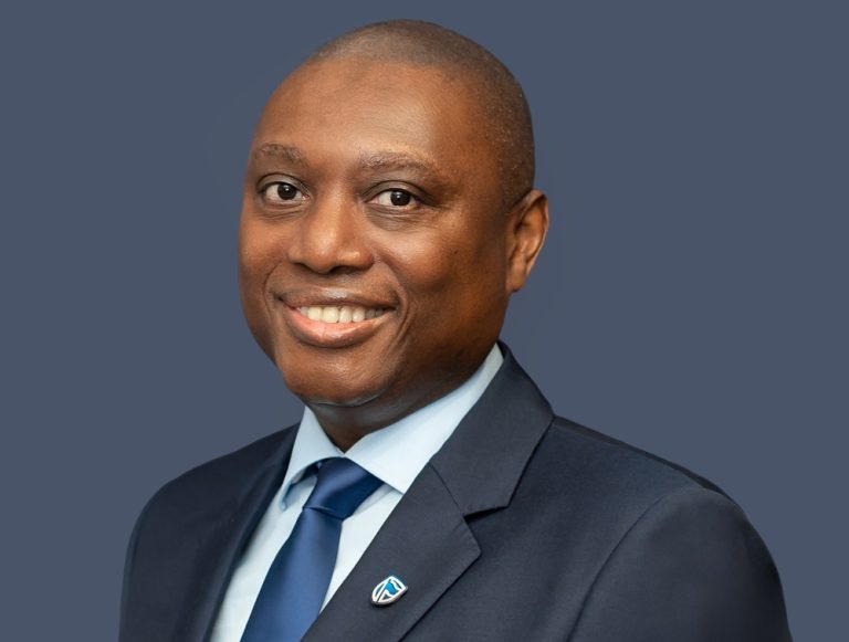 Smiling Black man in a navy suit and blue tie, wearing a lapel pin, against a blue-gray background (professional headshot).