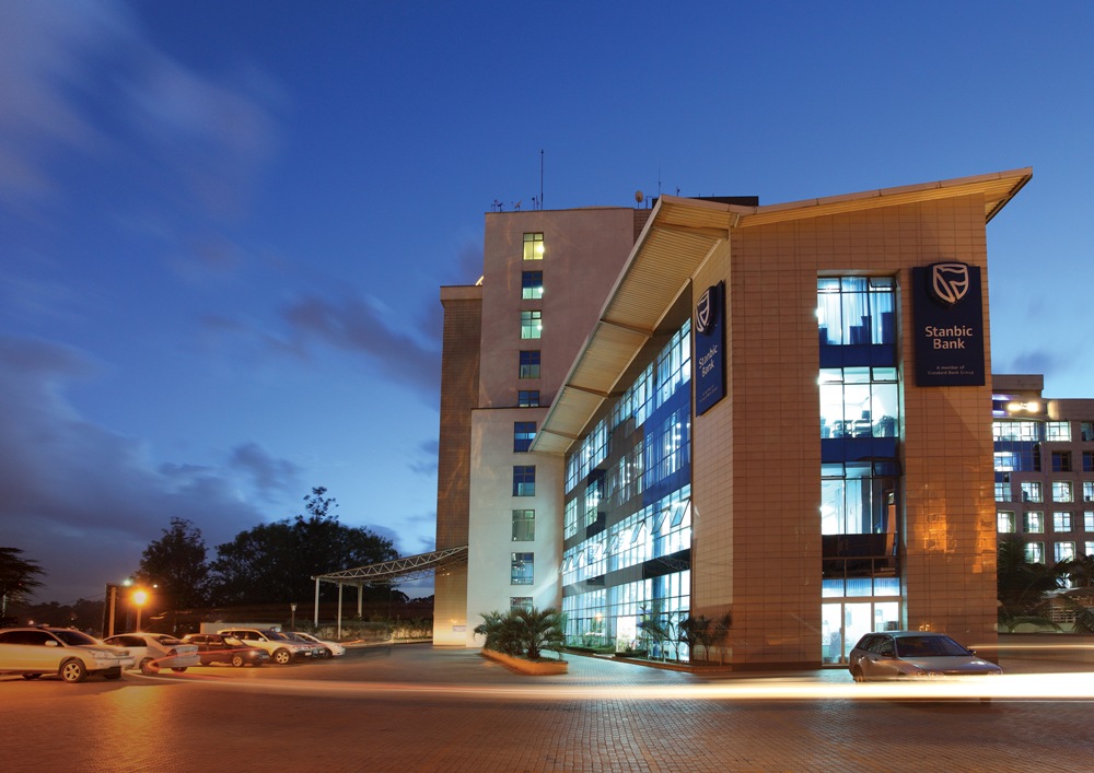 Exterior of Stanbic Bank building at dusk with glass facade and parked cars nearby on a paved lot, blue sky overhead.