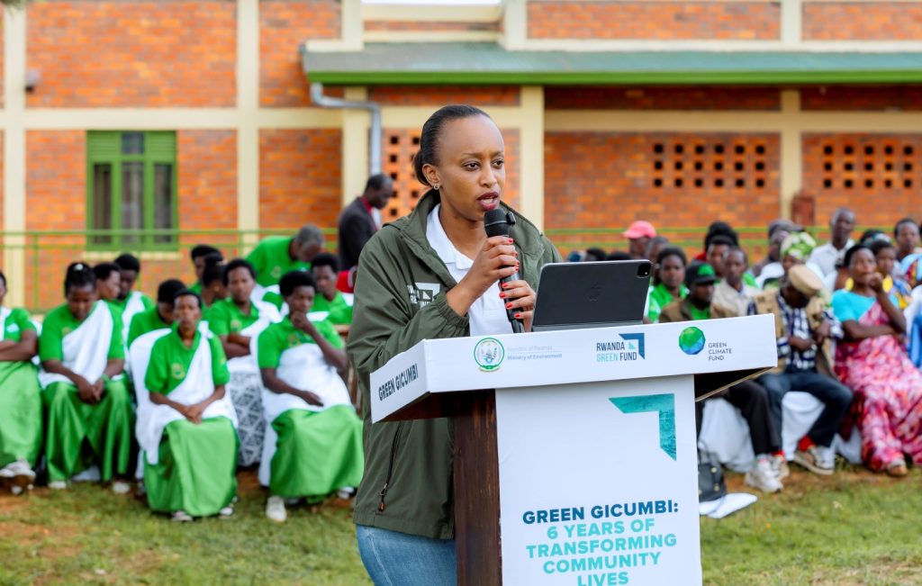 Woman speaks into a handheld mic at a podium during an outdoor event, with a green-themed audience behind her and banners mentioning Green Gicumbi and Rwanda Green Fund.