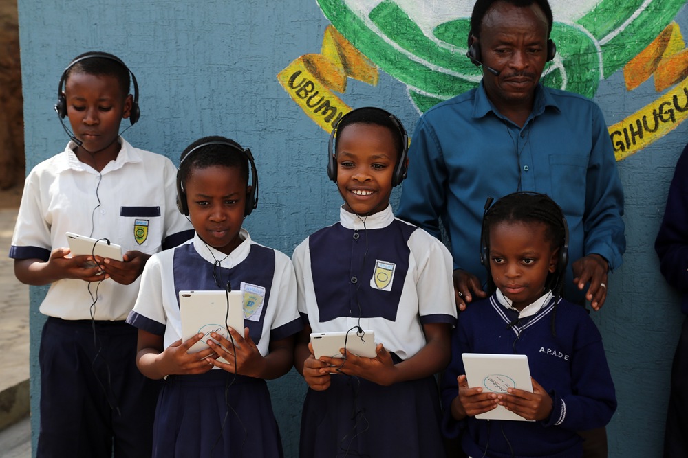 Four students in uniform with headsets hold tablets, standing in front of a mural while a man watches nearby.