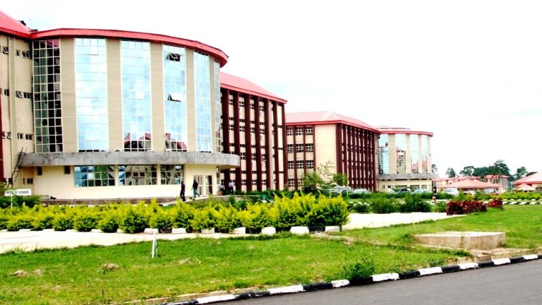 Modern campus building with curved glass facade and red roofs, surrounded by green landscaping and a paved road in foreground.