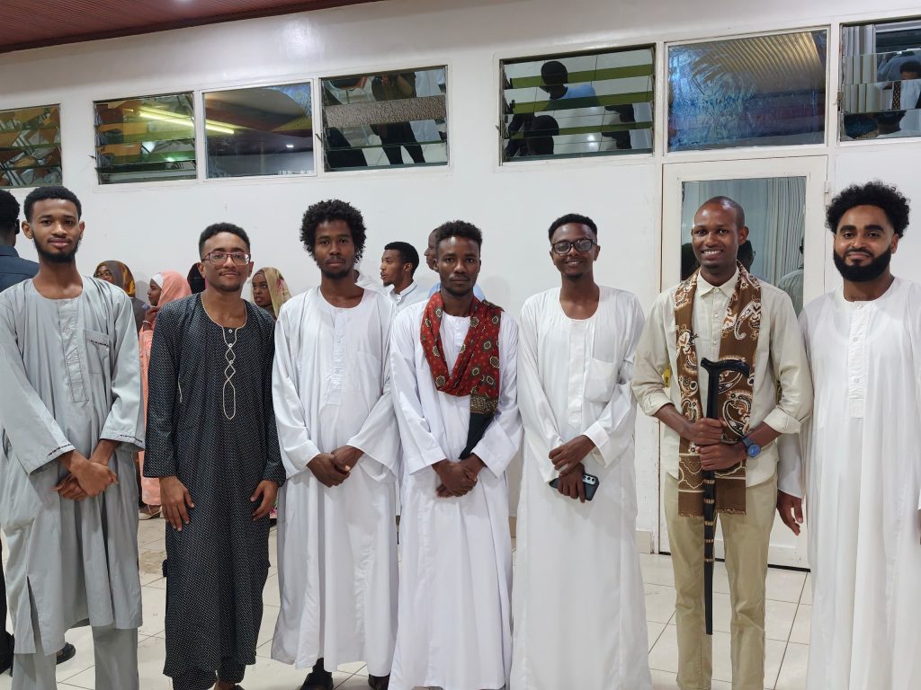 Line of men in white traditional robes posing for a group photo indoors, with others in background.